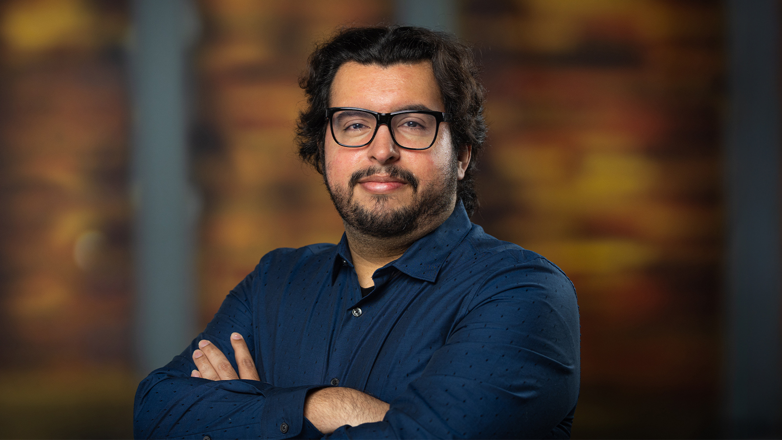 A man with glasses and a beard poses confidently with his arms folded against a warm, blurred background.