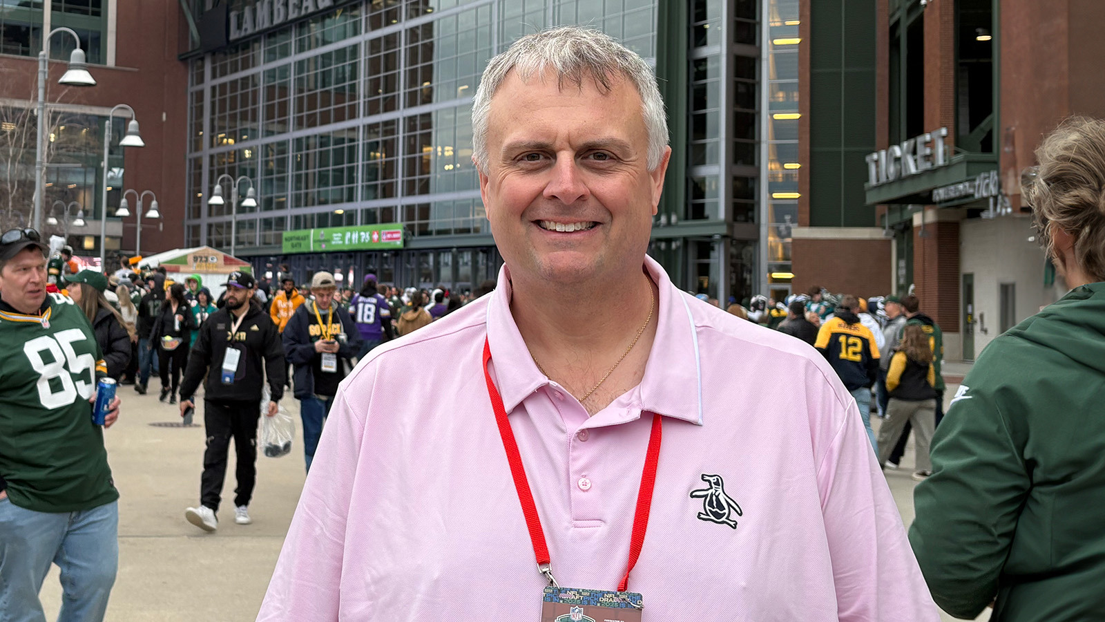 A man with short white hair and a pink polo shirt is seen smiling while standing in front of Lambeau Field in Green Bay, Wisconsin.