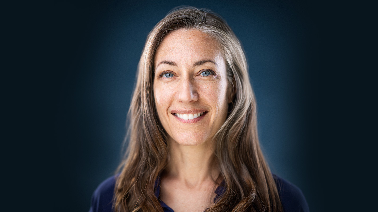 Kelly sits in front of a blue background wearing a blue blouse. She has blue eyes and is smiling.