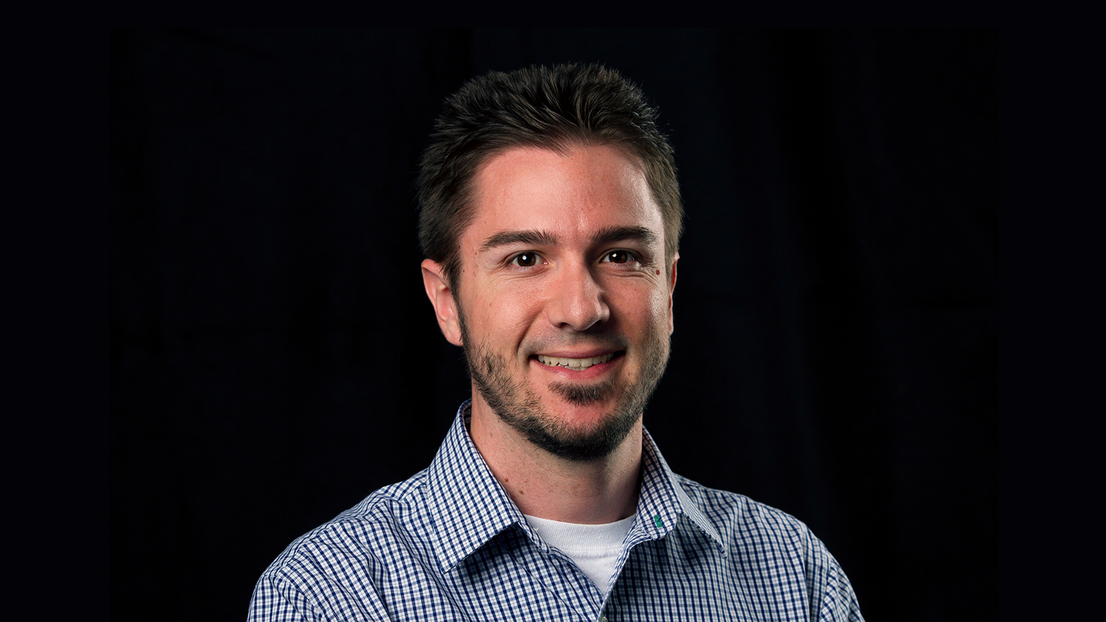 Matt is sitting in front of a black background wearing a blue plaid shirt. He is smiling.