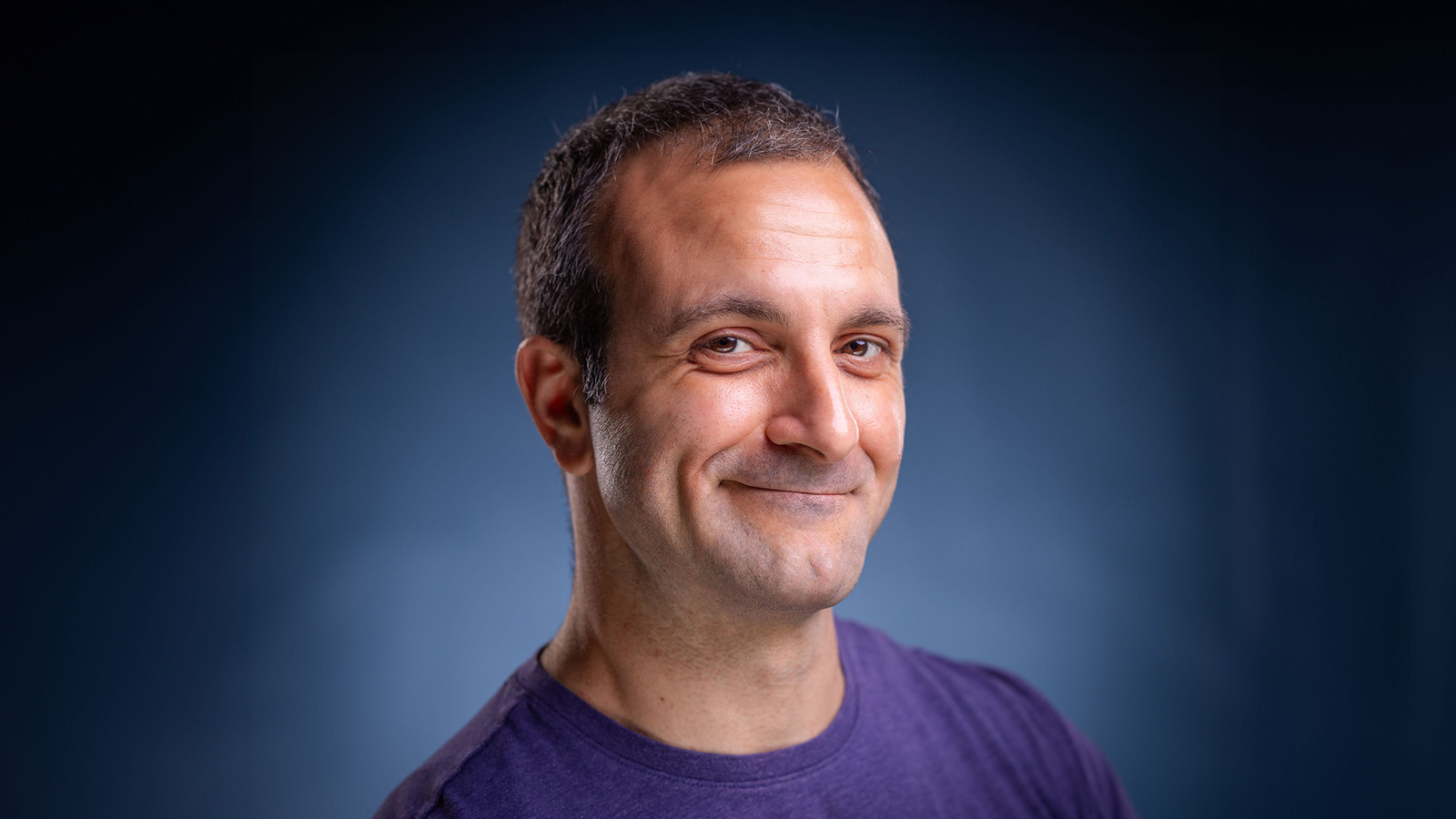 Michael Corinella smiles at the camera in front of a blue background. He is wearing a purple shirt.