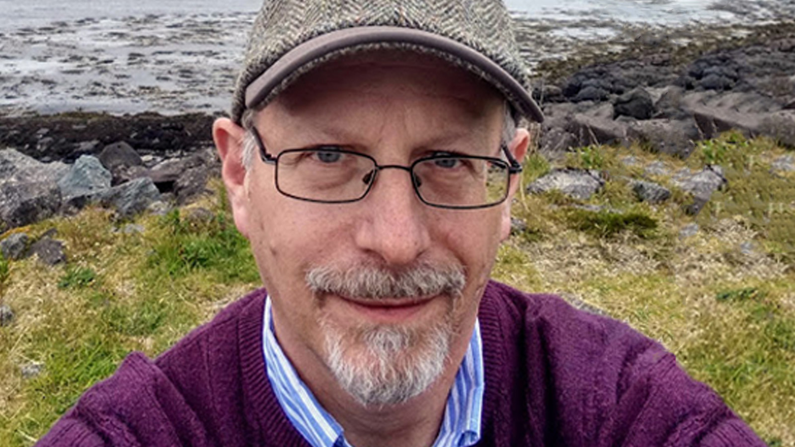 Peter Gordon stands outdoors in front of a river. He is wearing a purple sweater, a newsboy cap, and glasses.