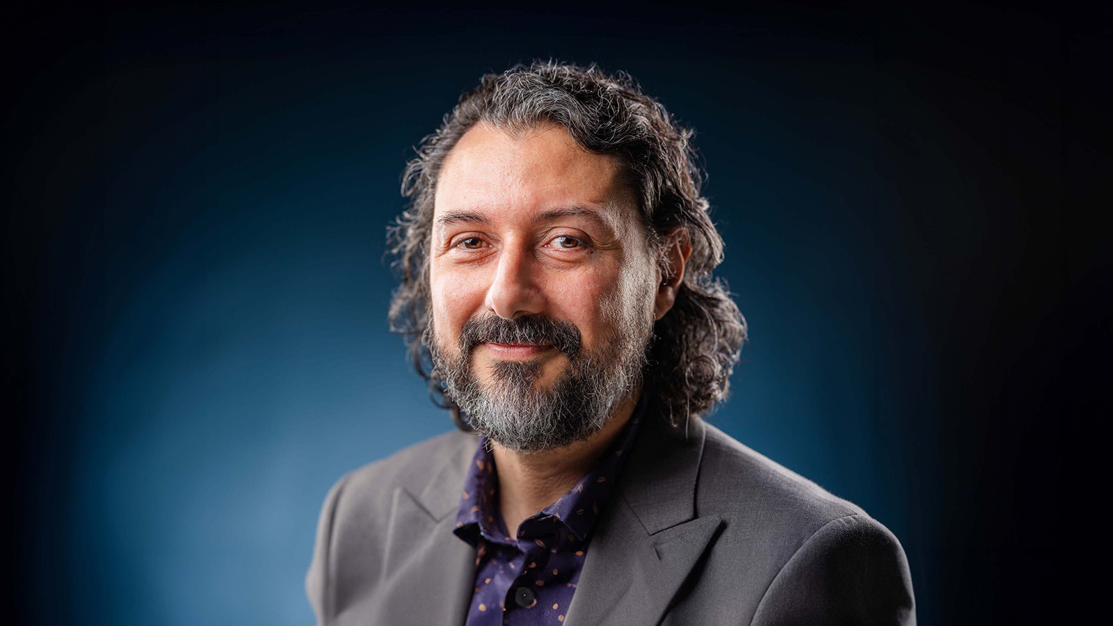 Plinio Pinto smiles at the camera. He has curly brown hair and a beard streaked with gray, and he wears a gray blazer.