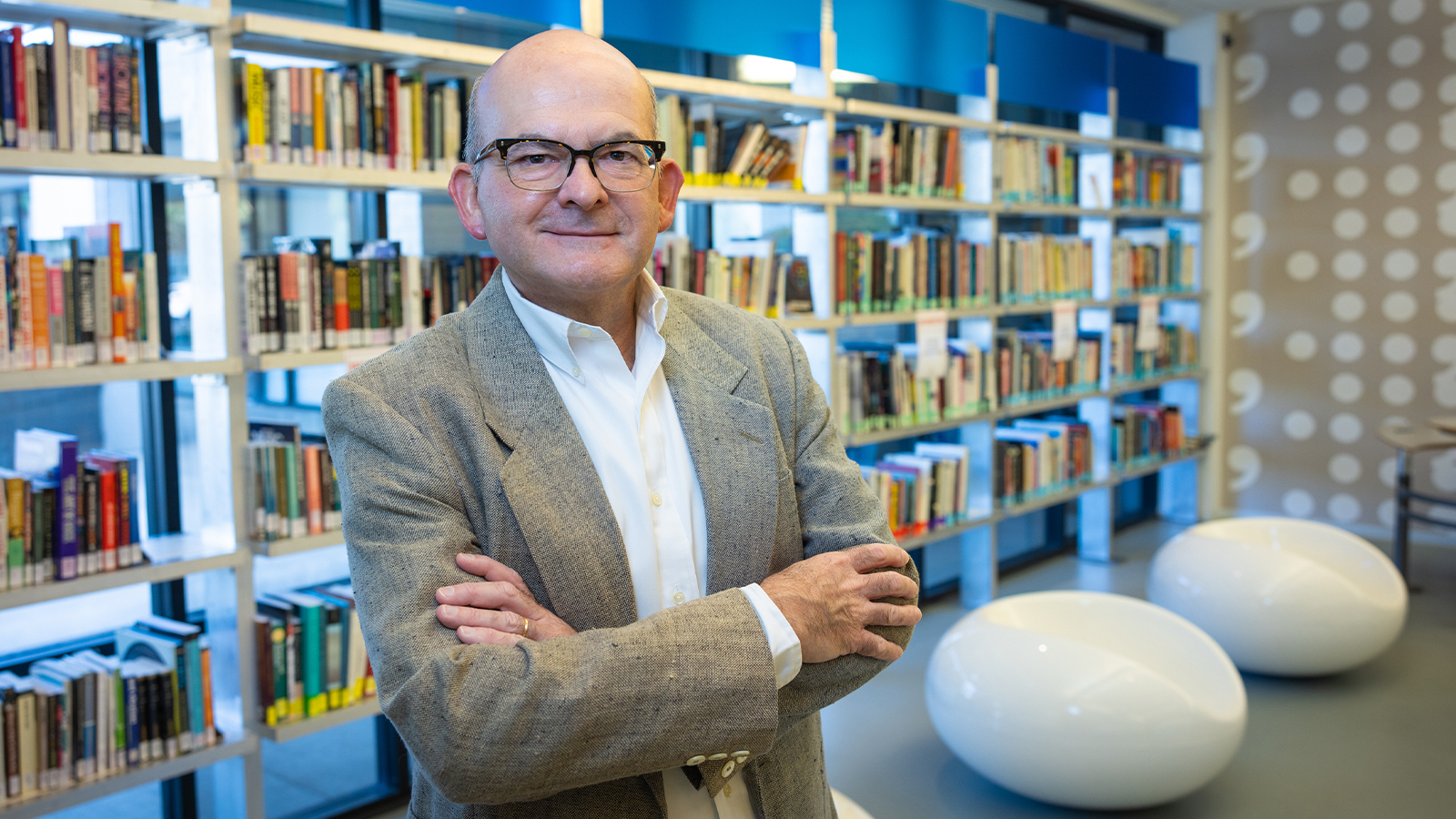 Rolando stands in a library with his arms crossed. He is smiling and wearing a suit.