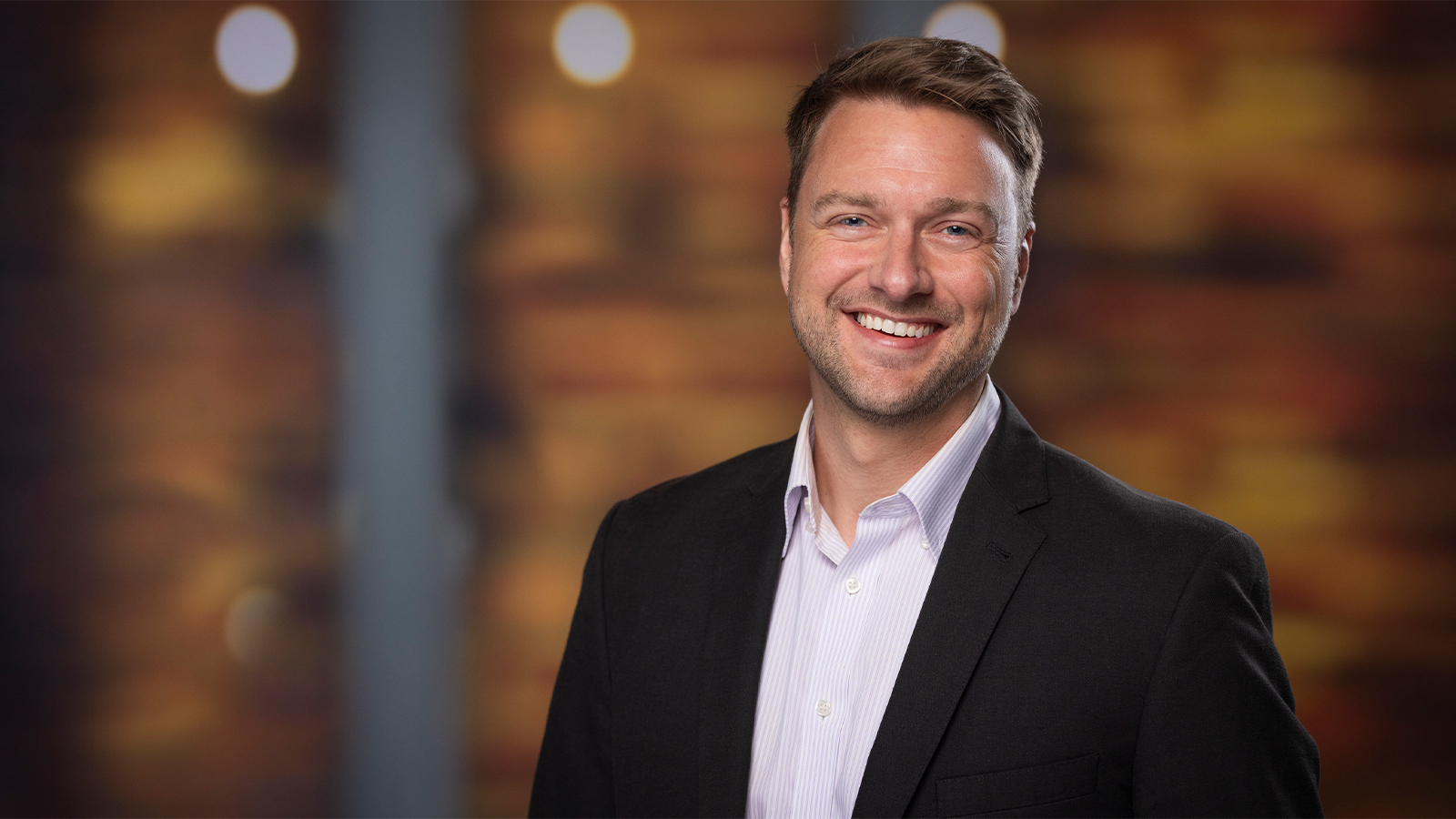 A smiling man in a black blazer and light dress shirt stands in front of a warmly lit, blurred background.