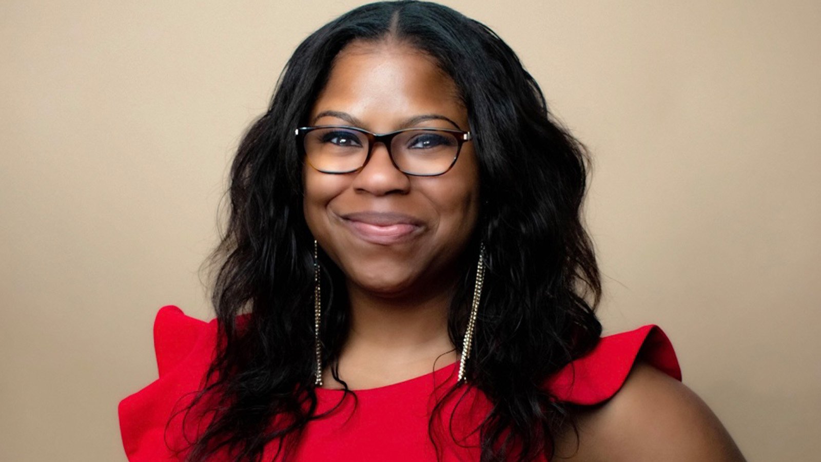 A woman with long, wavy black hair, glasses, gold earrings, and a red dress with ruffled sleeves smiles warmly.