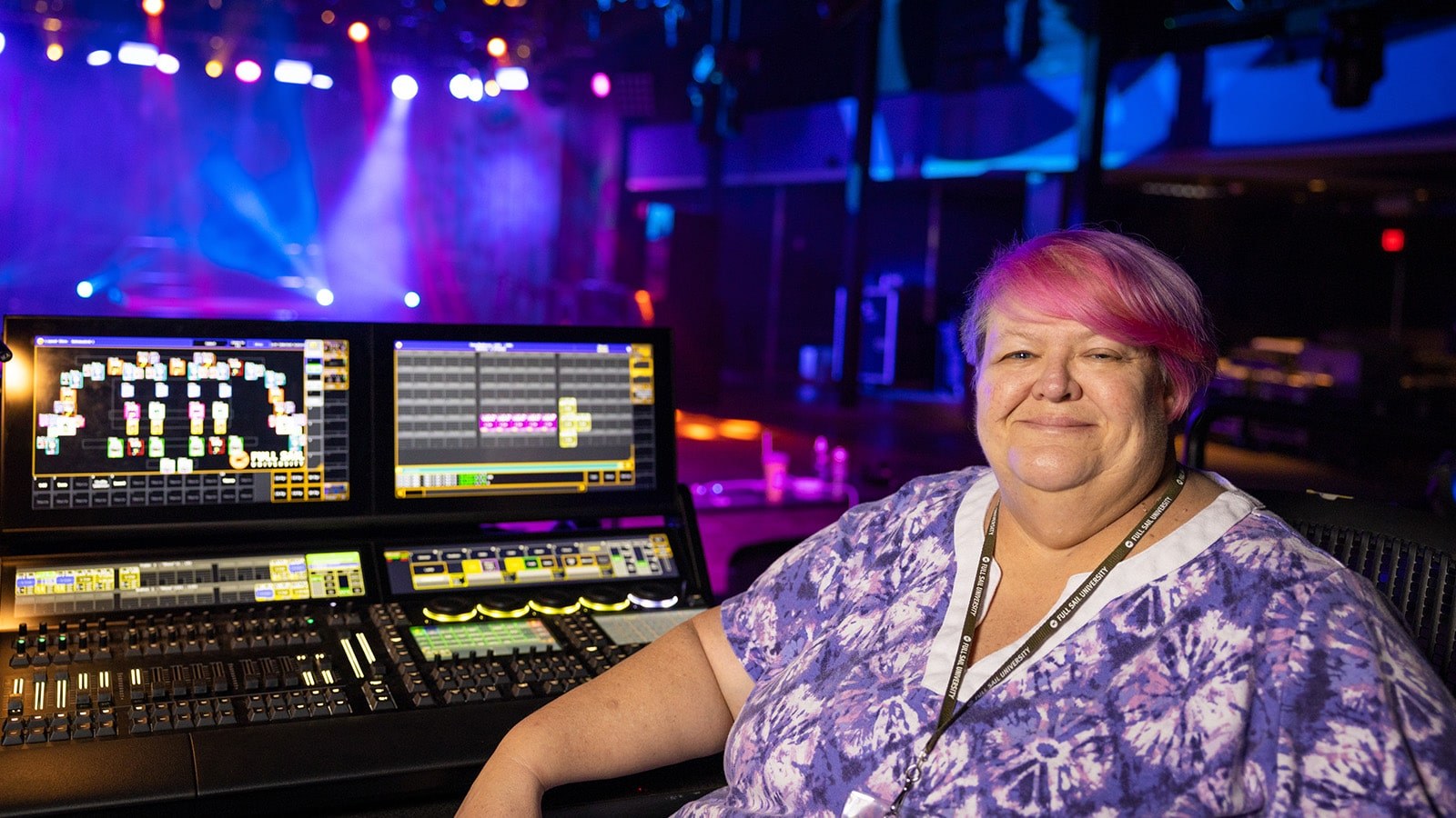 Susan Kelleher sits at a lighting console. The stage behind her is lit up with purple, blue, and white lights.