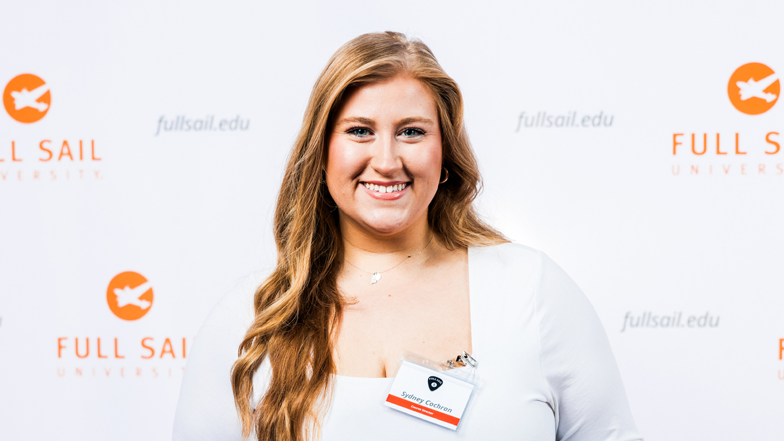A woman smiles while holding a Full Sail 5 year anniversary guitar pick in front of a Full Sail University step-and-repeat backdrop.