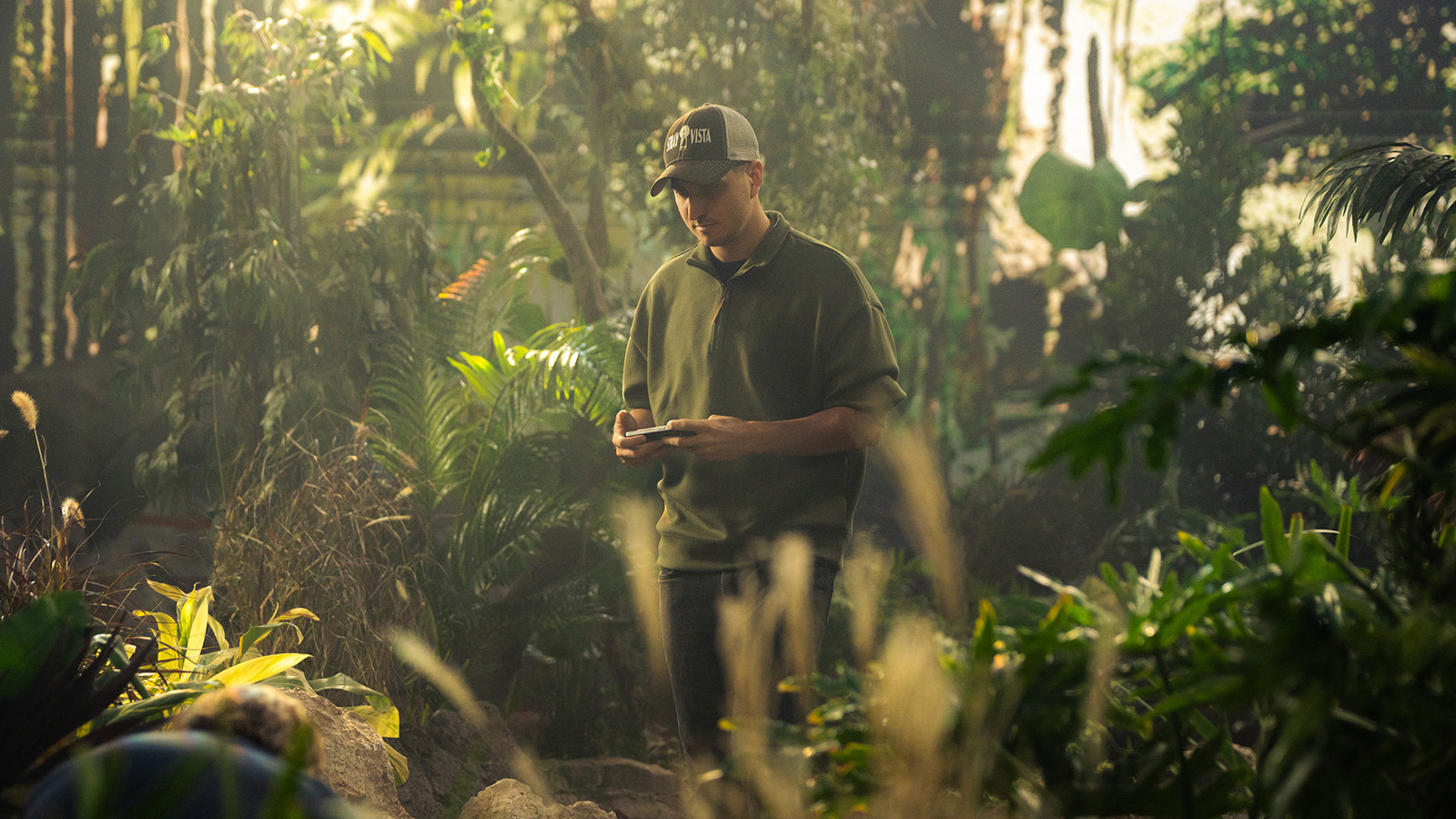 Nate Strayer stands on a film set filled with lush green trees and a digital forest background. He is wearing a baseball cap that says Stray Vista Studios.