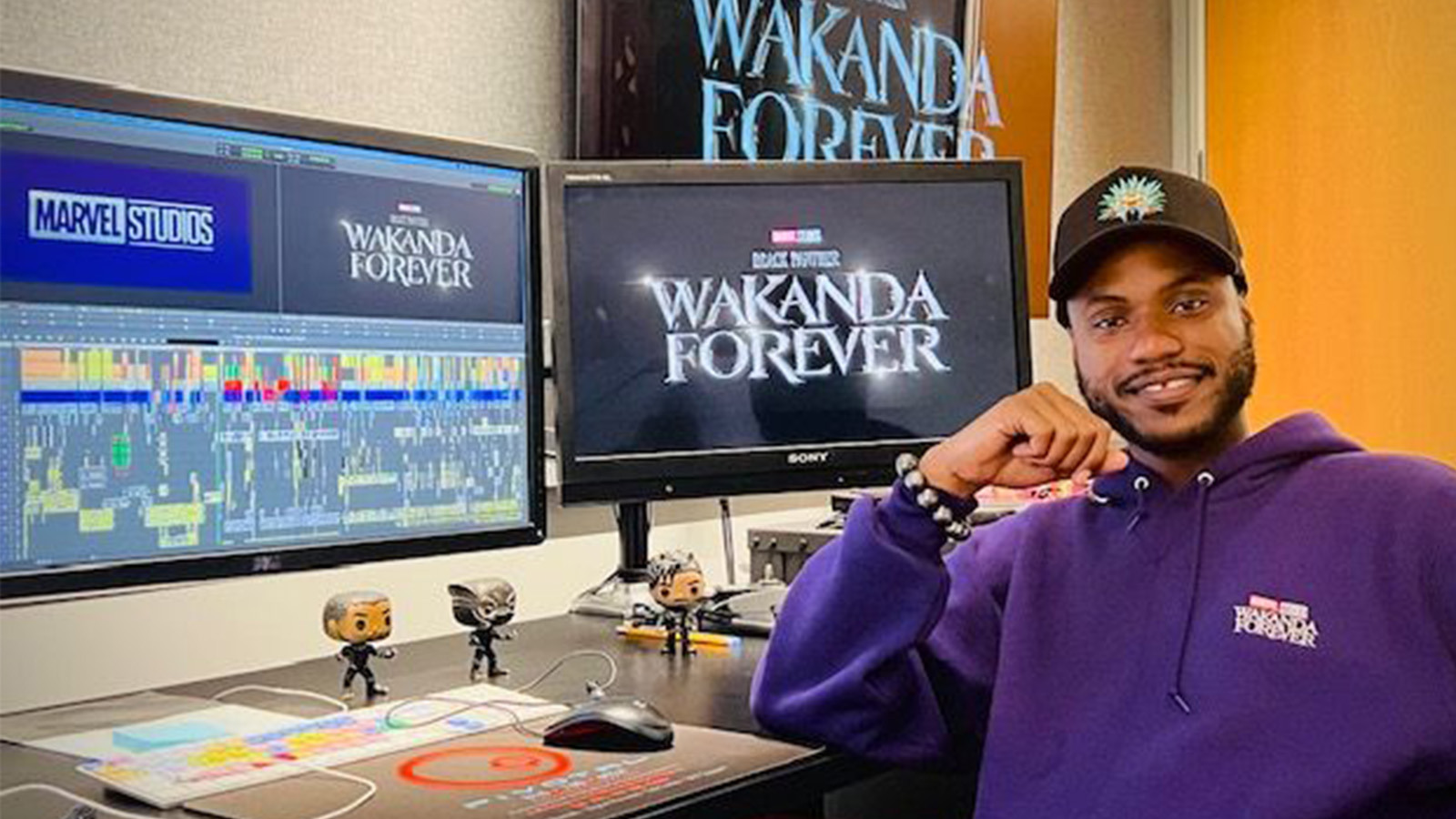 Alain sits at a desk with multiple monitors that read “Wakanda Forever” and “Marvel Studios.” He is wearing a purple hoodie.