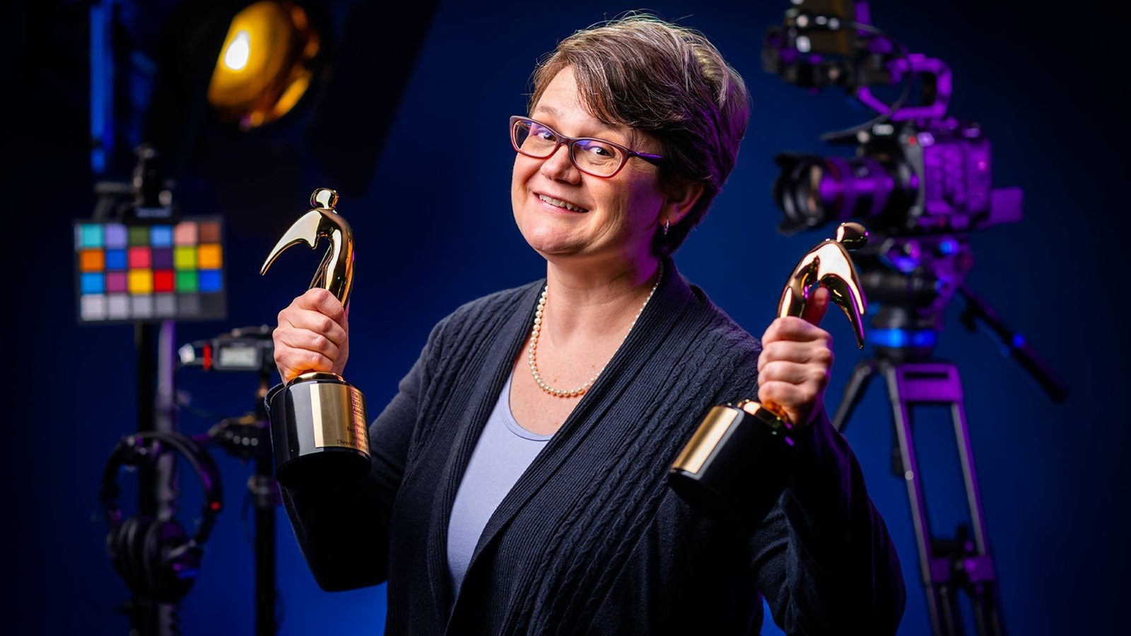 Jennie Jarvis stands in a studio with film cameras and stage lights. She is holding two Gold Telly statues.