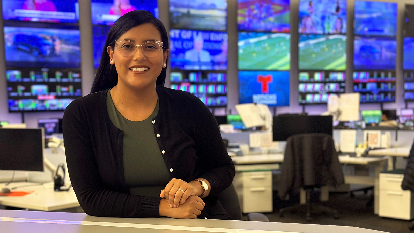 Priscilla sits at a desk in a live news television studio. She is wearing glasses and is smiling.