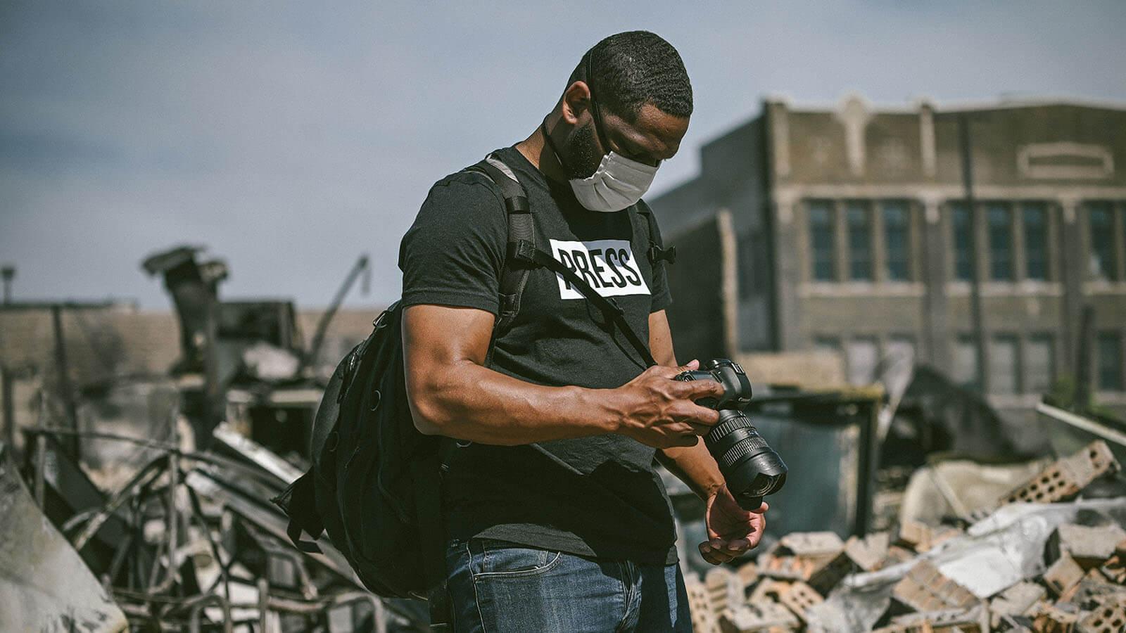 Grad E Mackey looking down at a DSLR camera while wearing a grey face mask, backpack, and black shirt with the word "press" in white, standing amongst a large amount of debris.