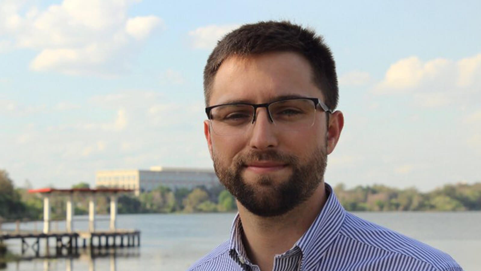 A man wearing glasses and a button-up shirt stands in front of a pier.