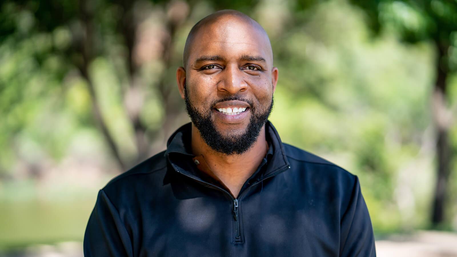 Full Sail grad Sidney Walker in the foreground smiling while wearing a black windbreaker with lush greenery behind him.