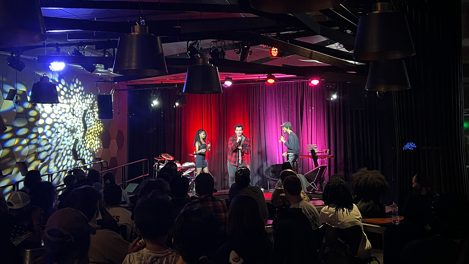 Three performers sing on a stage in front of an audience. Red and pink lights shine behind them.