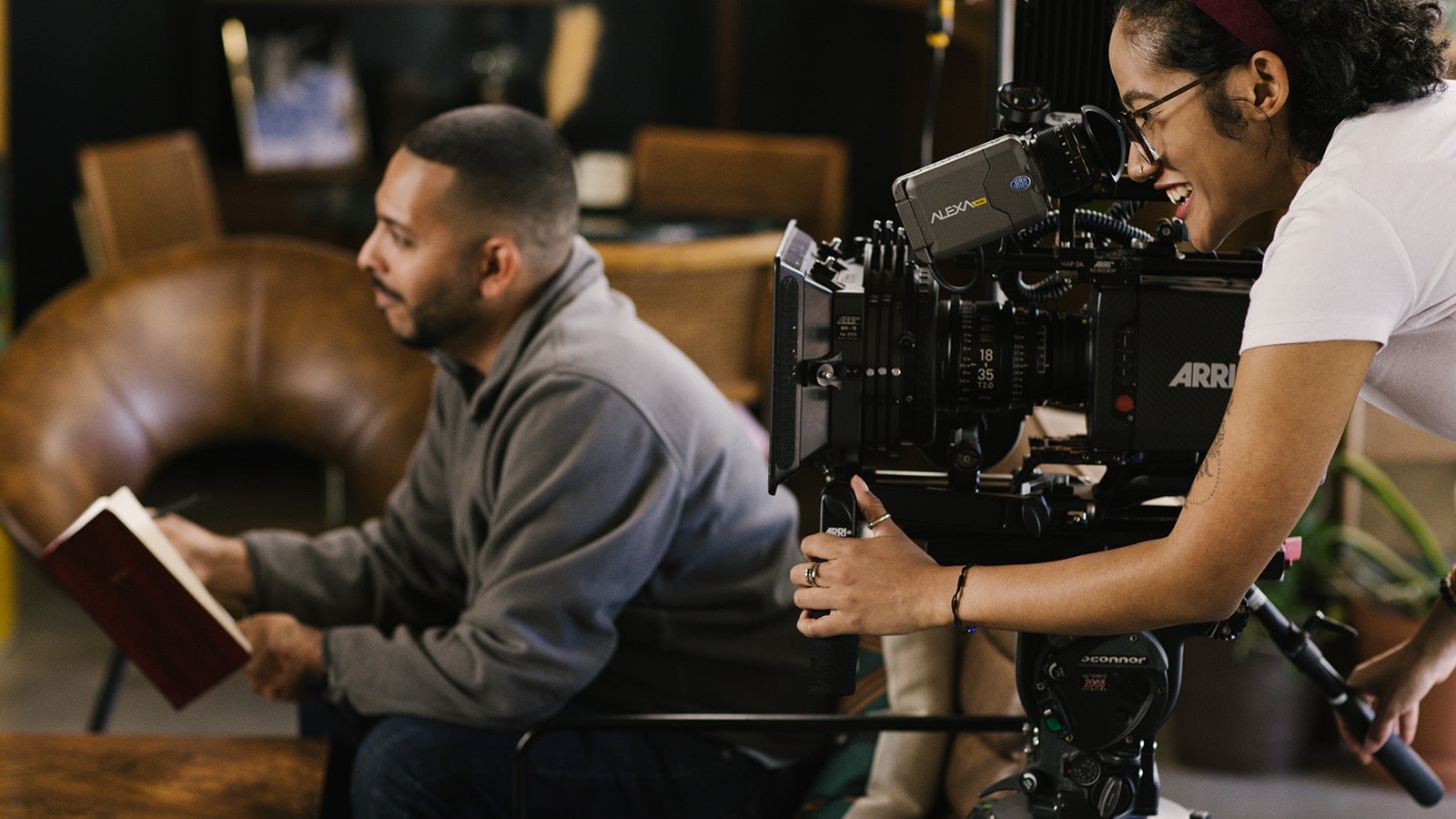 Behind the scenes on a student film set, one student sits in a chair with a notebook while another student works behind a film camera