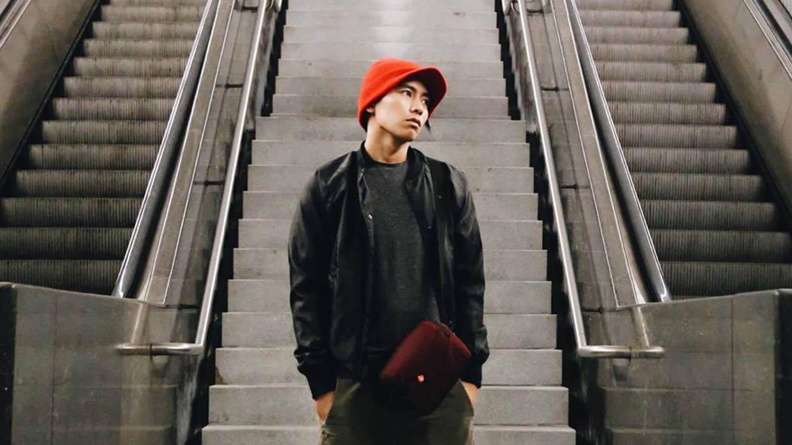 A young Asian man wearing a red knit cap, black jacket and shirt, green pants, and a burgundy bag stands between two escalators. His hands are in his pockets and he is looking off to the left.