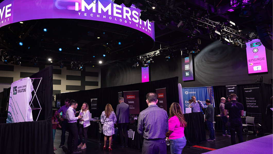 A large room with lots of booths set up, each from a different technology company. People in business-casual clothing are walking around and socializing.