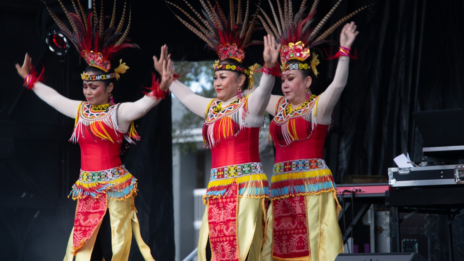 Three women dancing in colorful Indonesian feathered headdresses, yellow skirts, and beaded belts and collars.