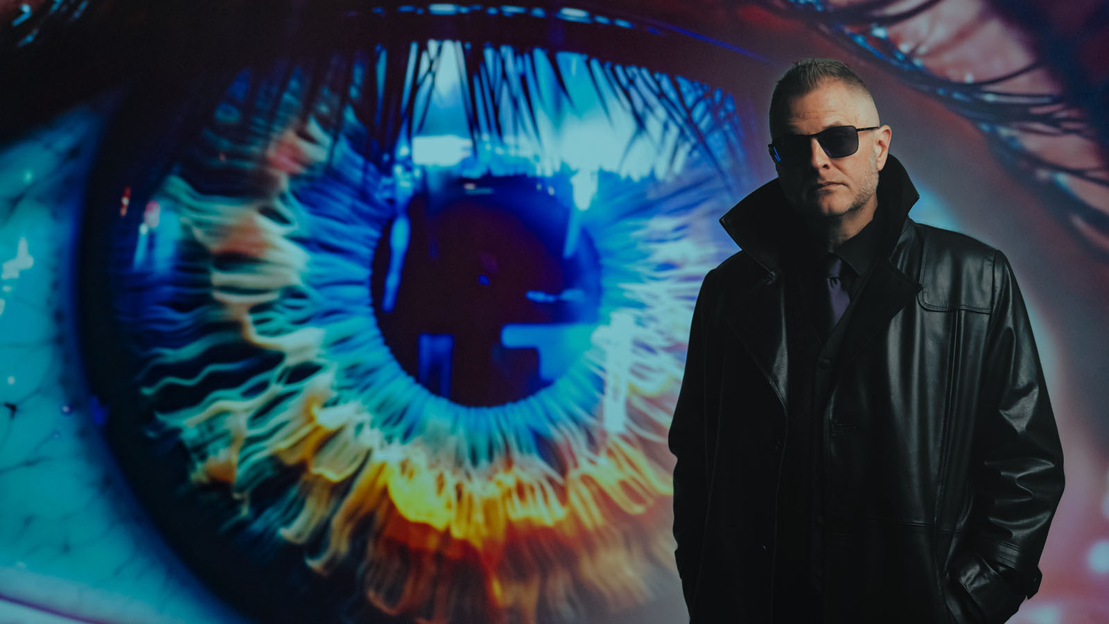 A man in a black leather coat and sunglasses stands before a giant, colorful close-up of an eye glowing with blue and orange light.