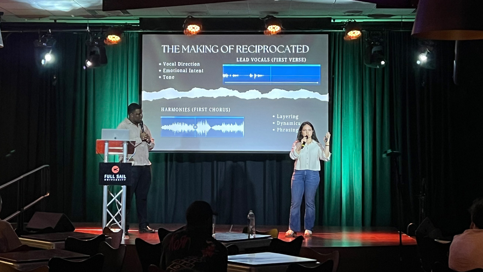 Tyonne Heyward and Ivana Loyola stand onstage in front of an audience. They share a slideshow presentation on the screen behind them.