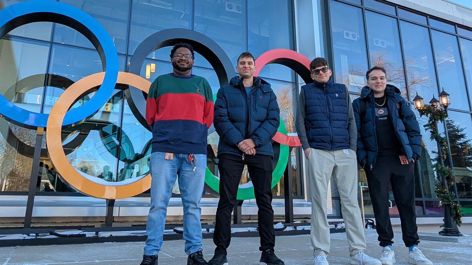 Aca Anderson, Johnathan Richardson, Jackson Faulknor, and Josh Minker stand in a row in front of the NBC Sports building with the colorful Olympic rings and a large sign displaying “NBC Sports.”