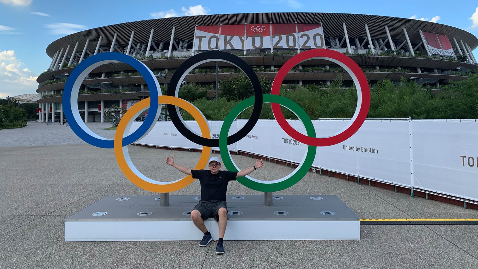 Grad Andrew Molina sits outdoors with his arms spread in front of a set of Olympic rings. There is a stadium with a banner for the 2020 Tokyo Olympics behind him.