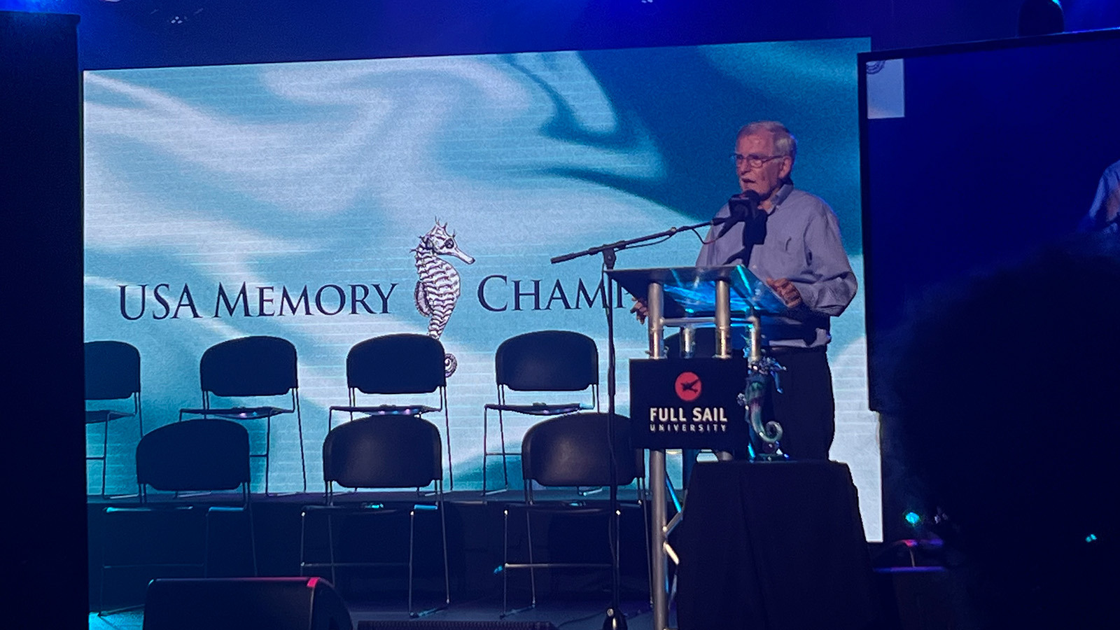 A man wearing glasses and a collared shirt speaks at a podium on a stage. A screen that says USA Memory Championship is behind him.