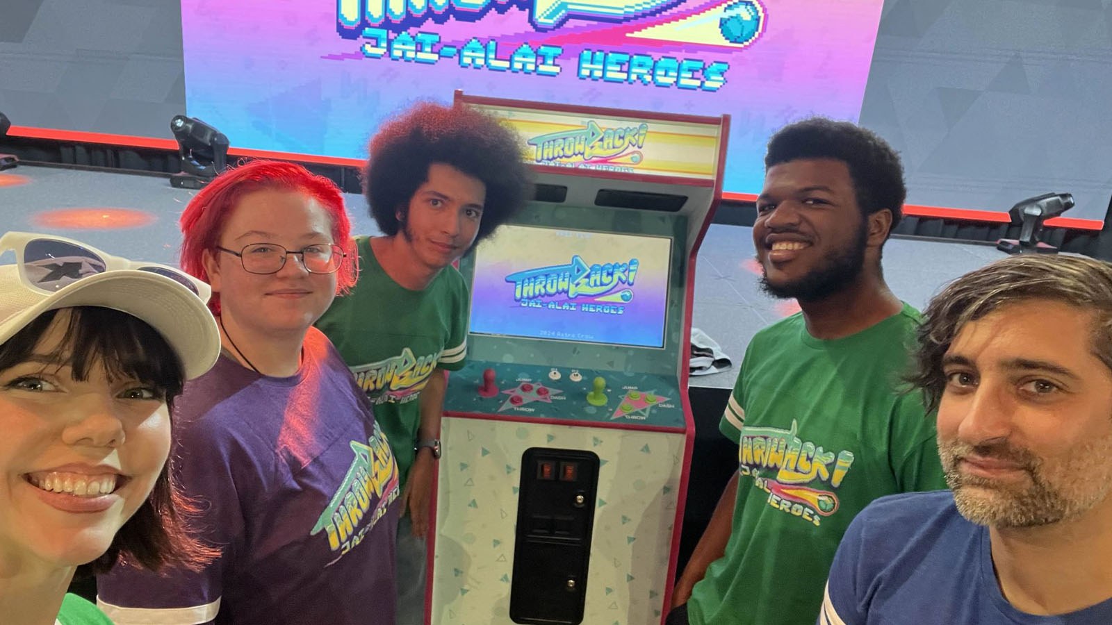 Caris Baker stands with four students in front of an arcade game machine that says Throwback! Jai-Alai Heroes on the screen.