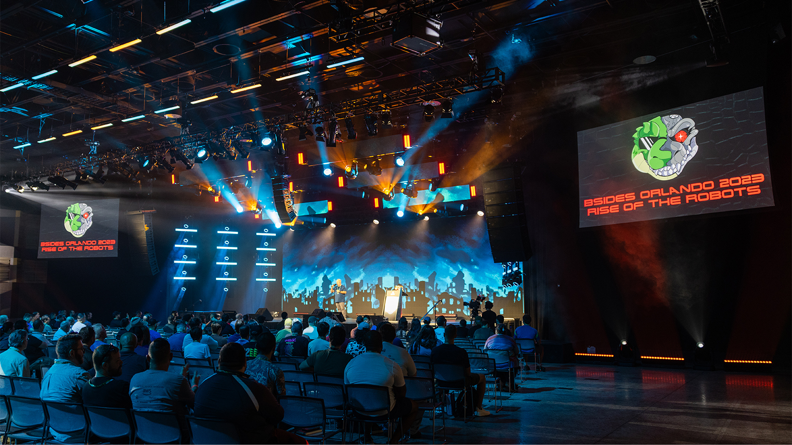 Attendees sit in the Full Sail Live venue. Screens with a lizard robot logo and the words BSides Orlando 2023: Rise of the Robots are on either side of the stage.