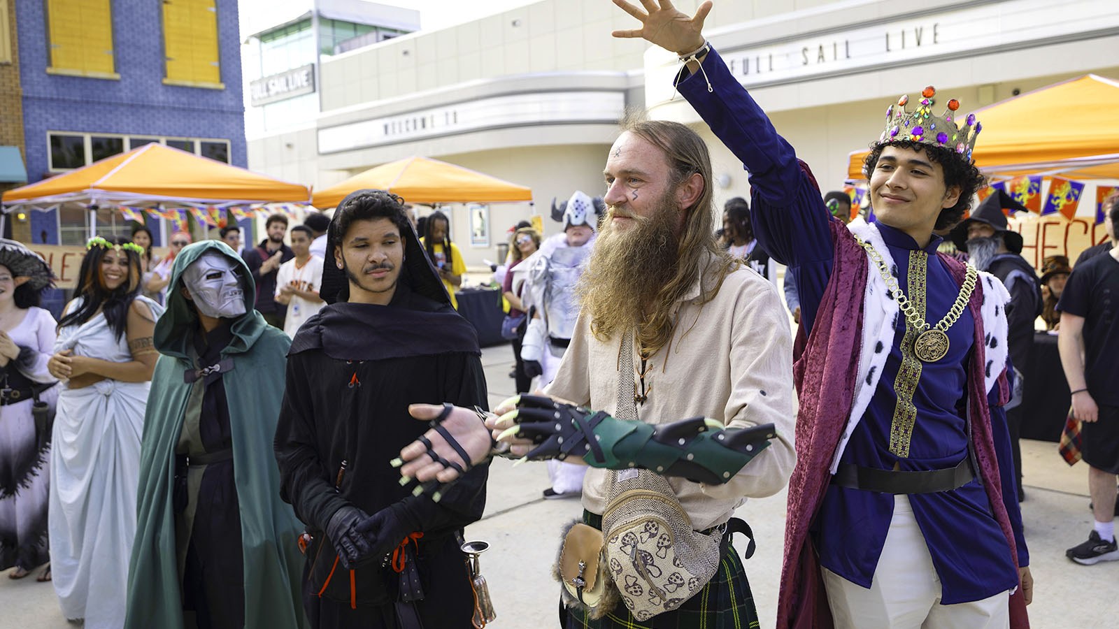Several students dressed in costumes including a mushroom, a goddess, a viking, and a king stand in a line. Orange tents surround the crowd of students.