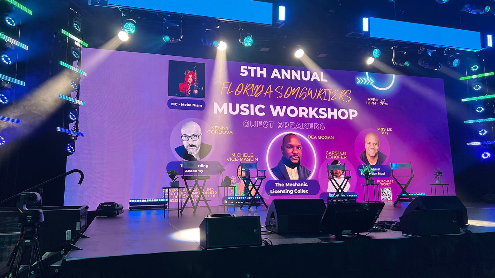 A large screen reads "5th Annual Songwriters Music Workshop" against a purple backdrop. The screen sits atop a brightly lit, empty stage.