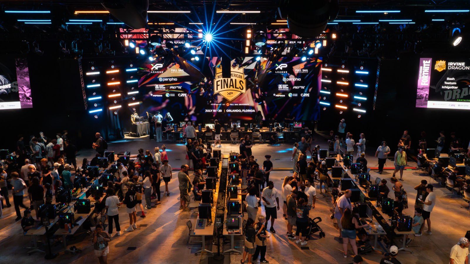 Four rows of 12 gaming consoles each fill a large room. Bystanders watch as competitors play various games at their stations. A large screen displays an XP League North American Finals graphic.