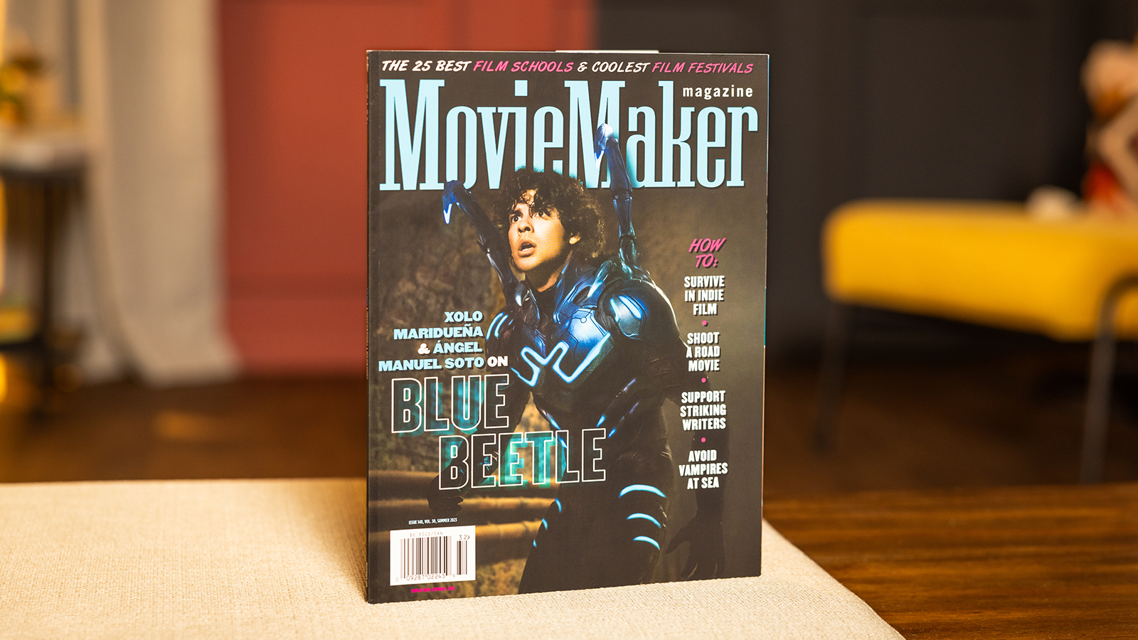 A copy of MovieMaker Magazine sits on a table. An actor in a Blue Beetle costume is on the cover. The headline “The 25 Best Film Schools & Coolest Film Festivals” runs across the top.