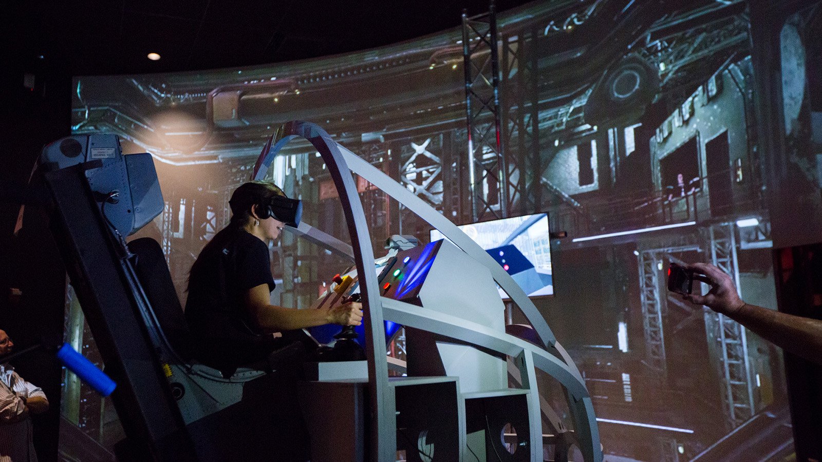 A student wearing a VR headset sits in a space shuttle landing simulator. A large screen displaying a warehouse is in front of them.