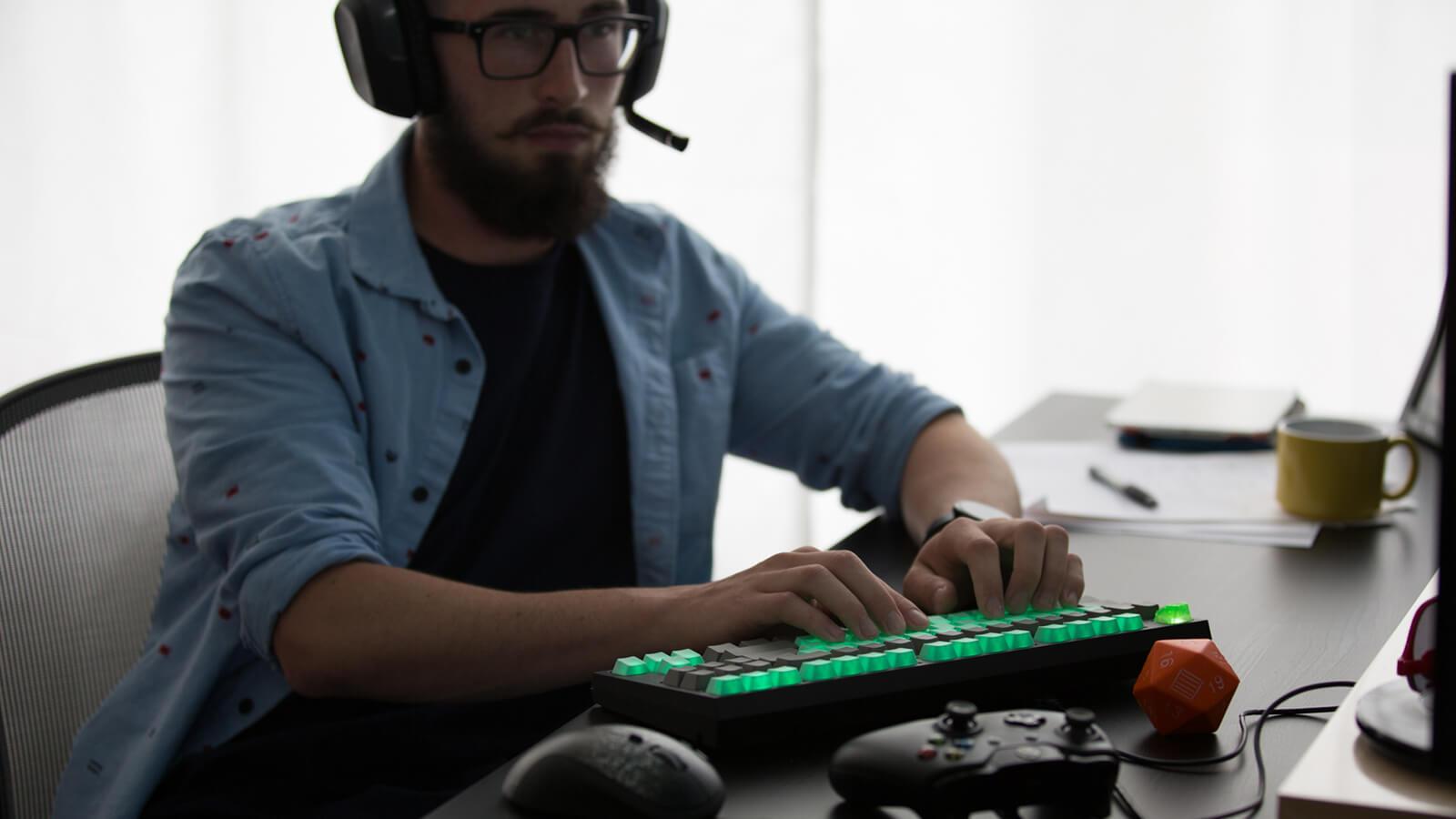 A man wearing a gaming headset sits at a desk with a gaming keyboard, an Xbox controller, and a Dungeons & Dragons die.