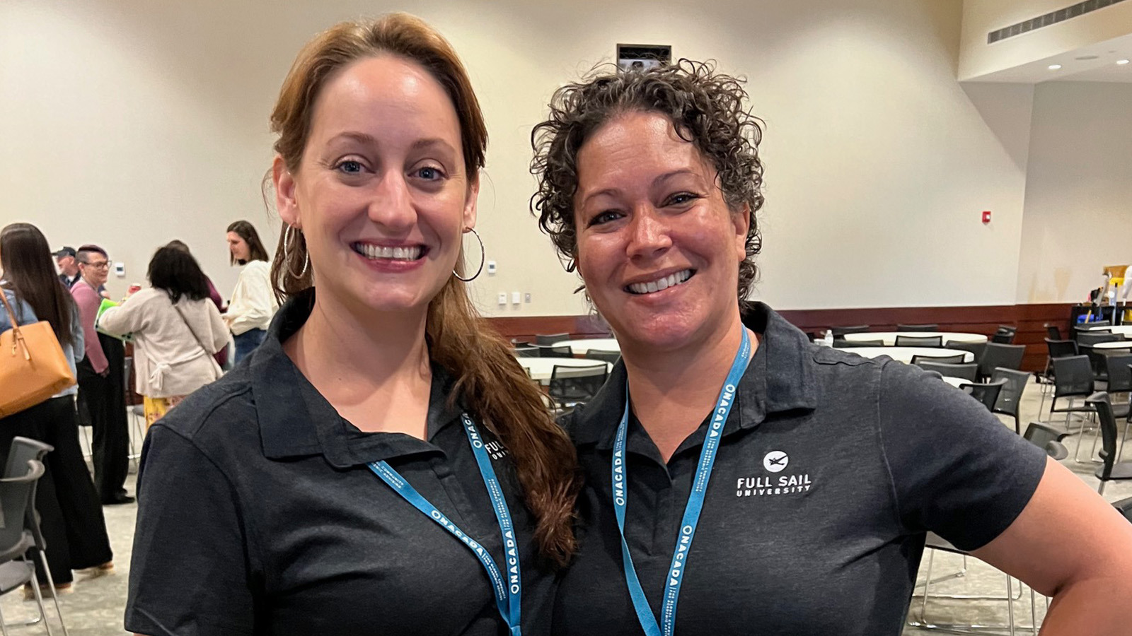 Michelle Moreno and Stacie Aldrich smiling and standing side by side wearing blue lanyards with conference badges and black Full Sail polo shirts.