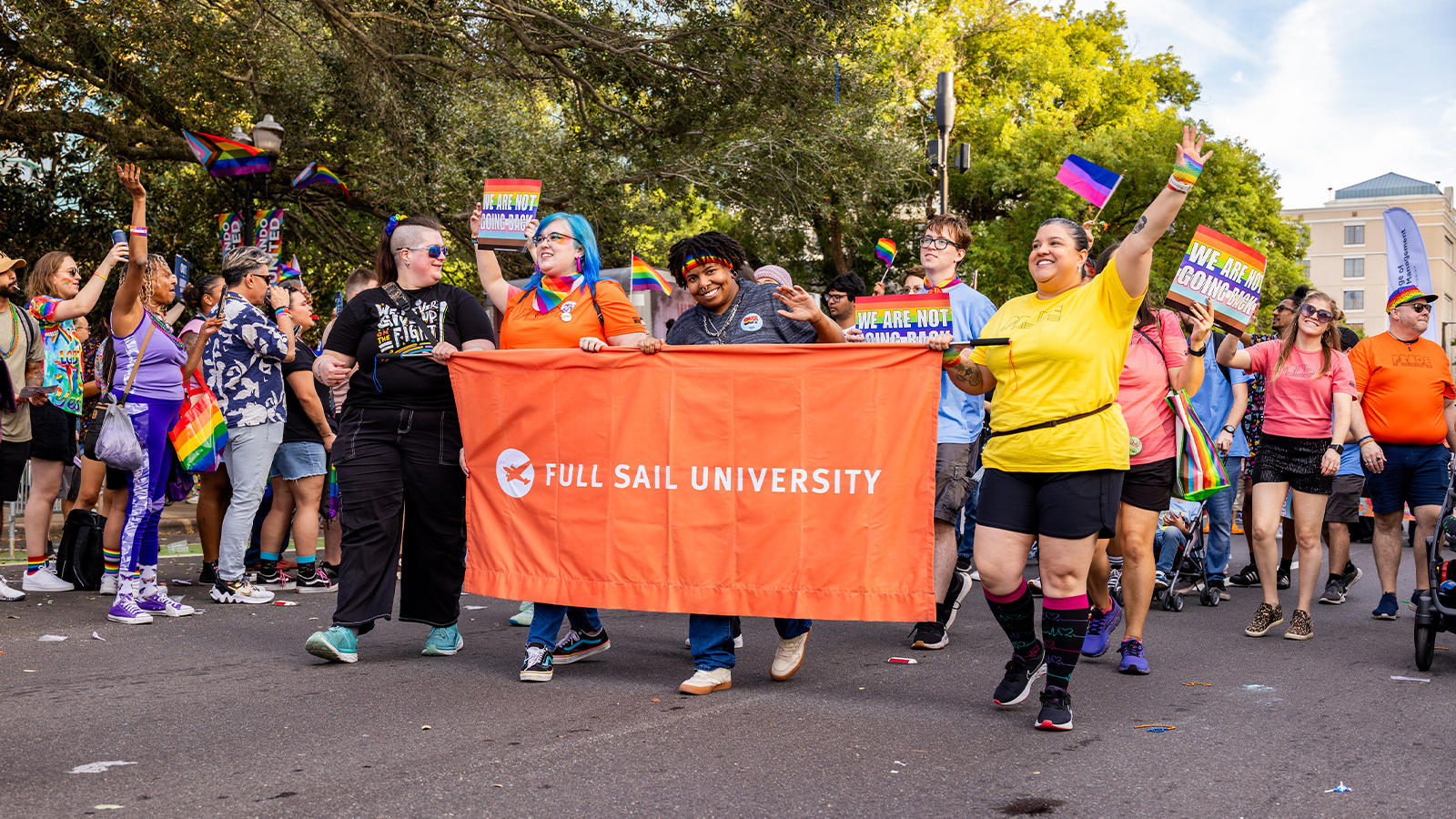 Full Sail students holding a Full Sail University banner march during the Pride Parade. A crowd holding rainbow signs and flags walks behind them.