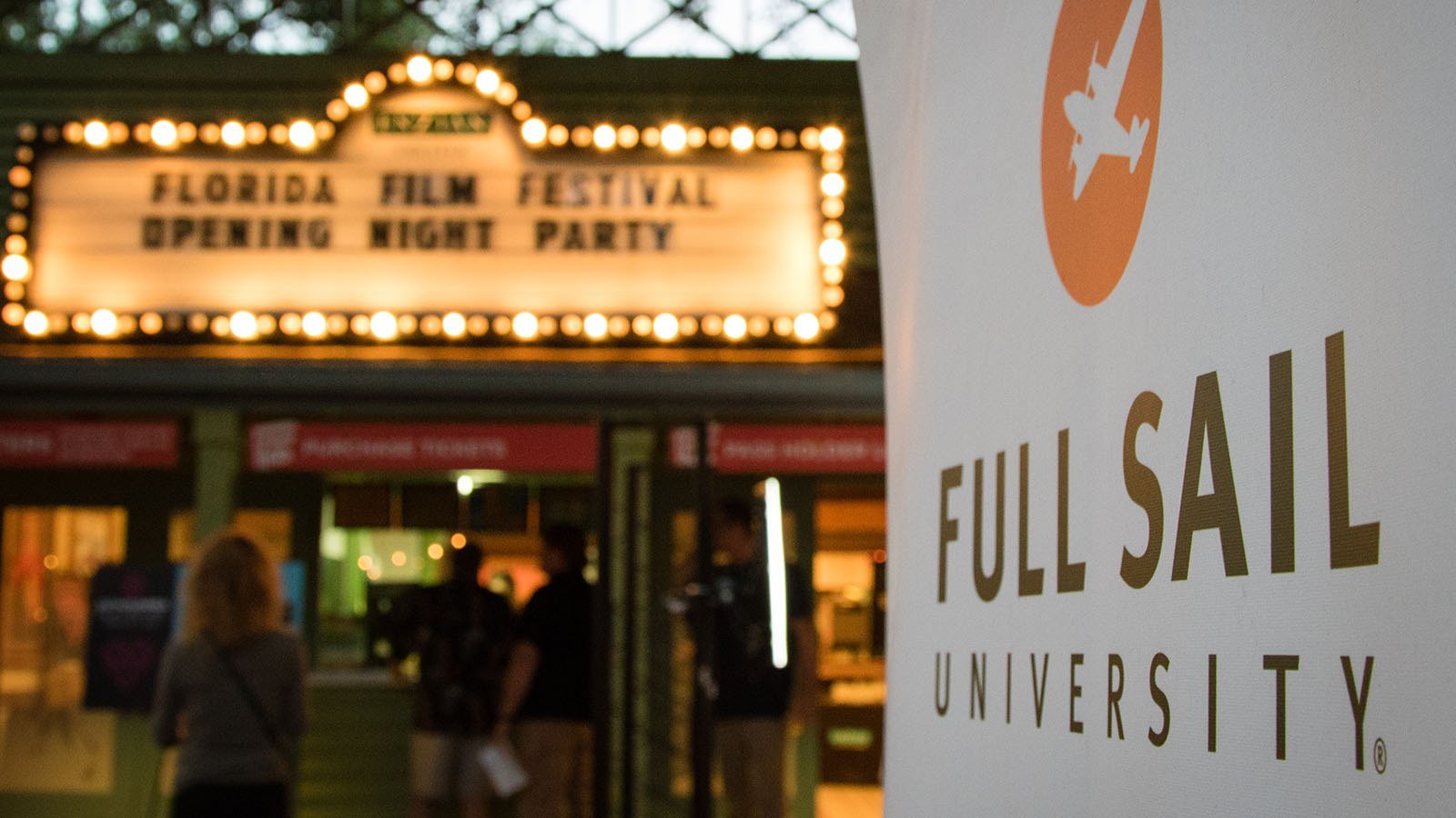 A Full Sail University banner is in focus in the foreground while the Enzian's lit-up marquee for the Florida Film Festival Opening Night Party appears in the background.