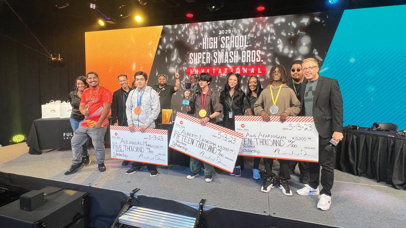 Techfest tournament winners holding large scholarship checks on stage alongside Full Sail staff members in the Fortress.