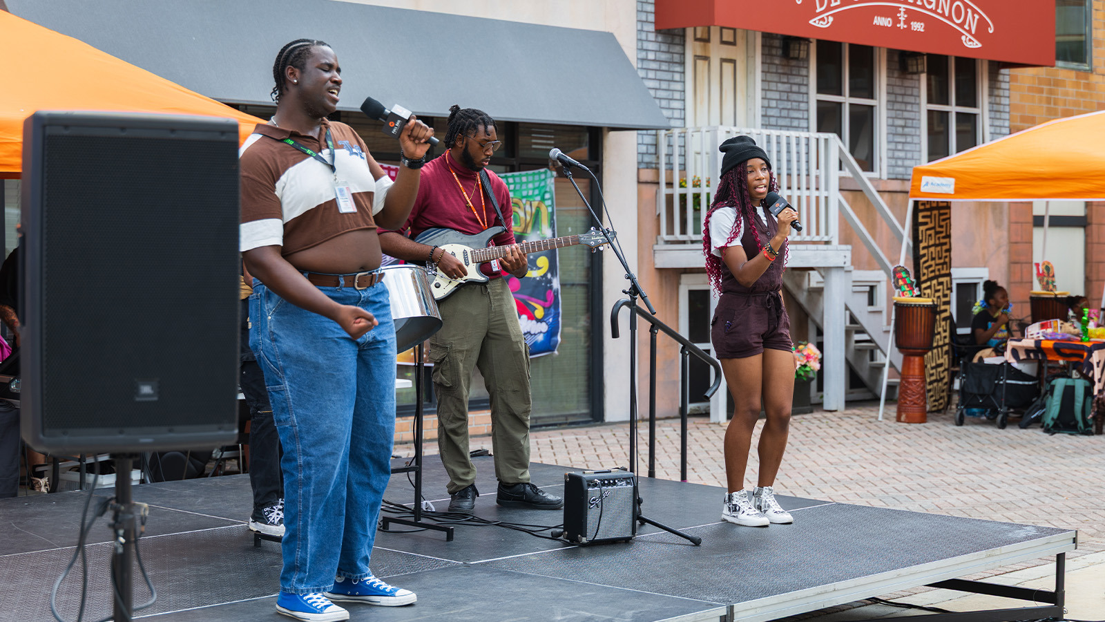 Three performers stand onstage. Two are holding microphones and singing, and one is playing an electric guitar.