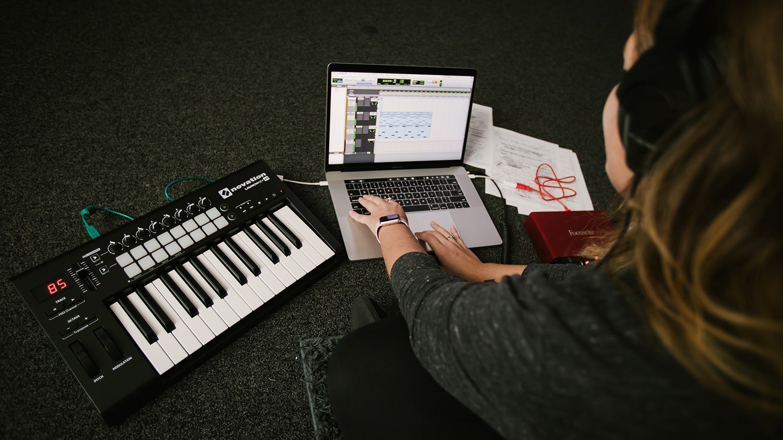 A student wearing headphones sits on the floor with a laptop and electronic keyboard. Music editing software is on the laptop screen.