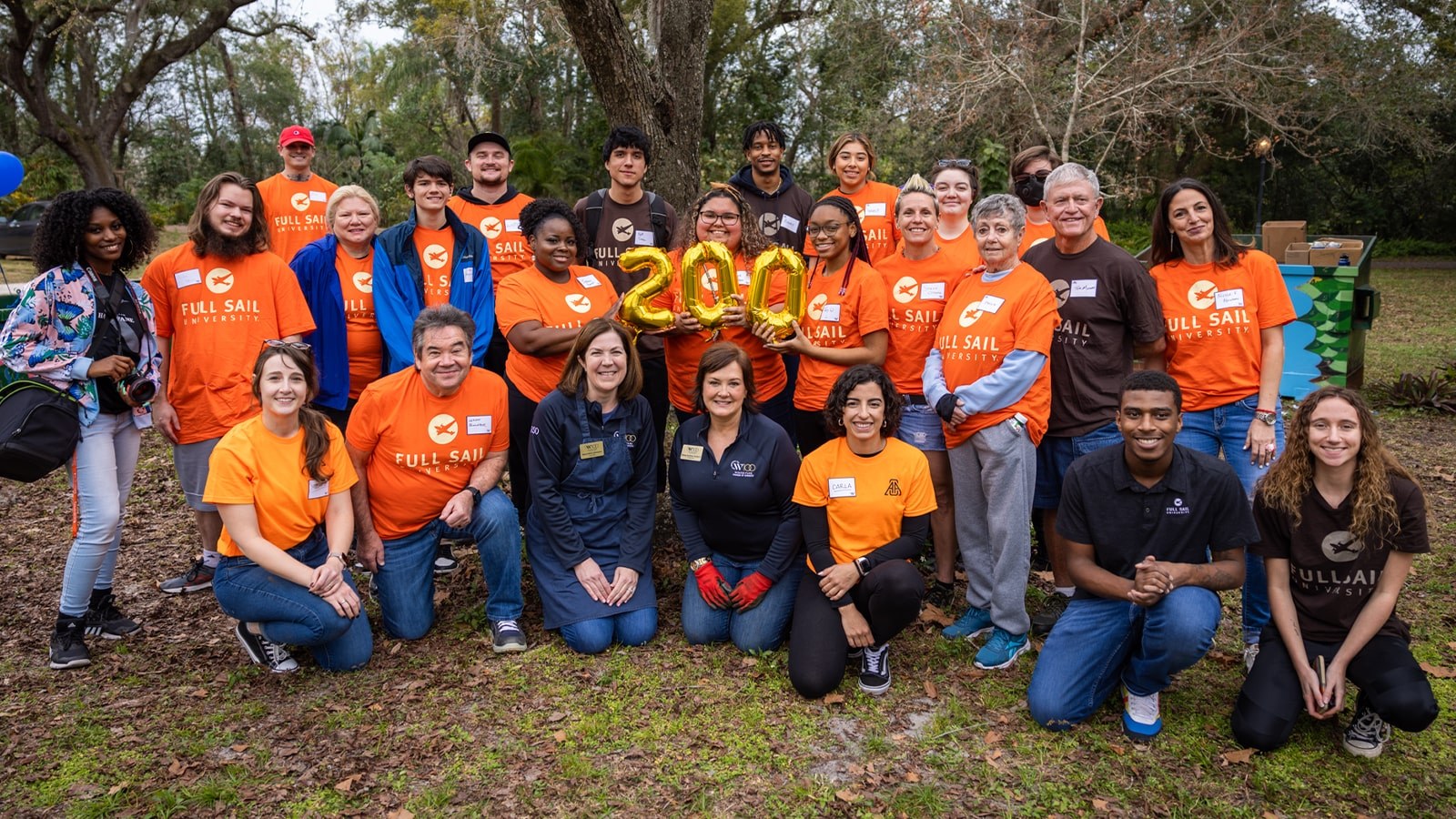 A group photo of students and staff wearing bright orange Full Sail University t-shirts. They are also holding golden balloons that spell 200.
