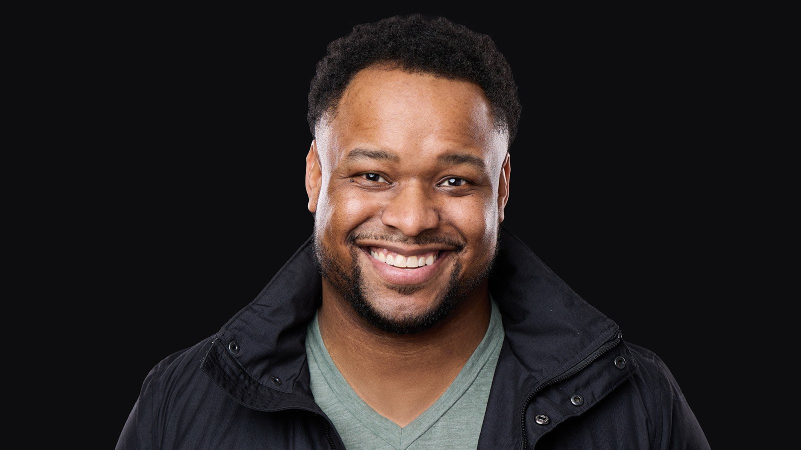 James Lewis smiles at the camera. He is wearing a black jacket and green v-neck t-shirt.