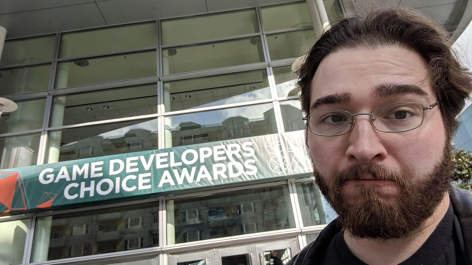 Ryan Miller pictured from above the shoulders in a black leather jacket with silver wireframe glasses in front of a sign reading “Game Developers Choice Awards.”