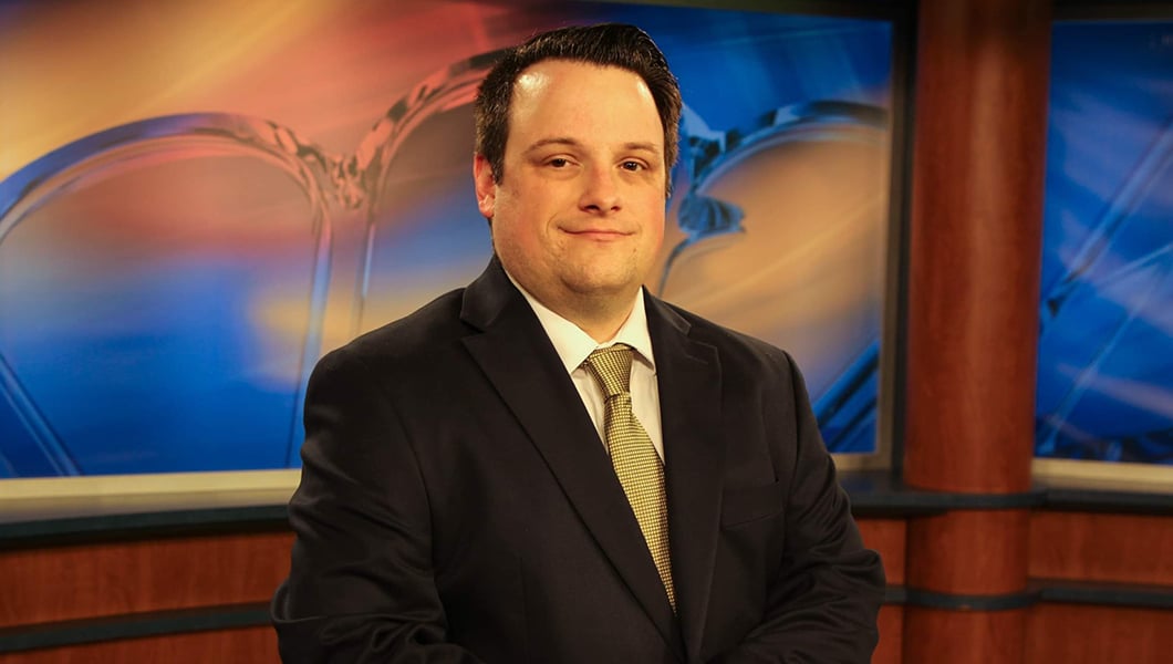 Dan Hanson sits at a news desk in a television studio. He is smiling and wearing a black suit with a yellow tie.