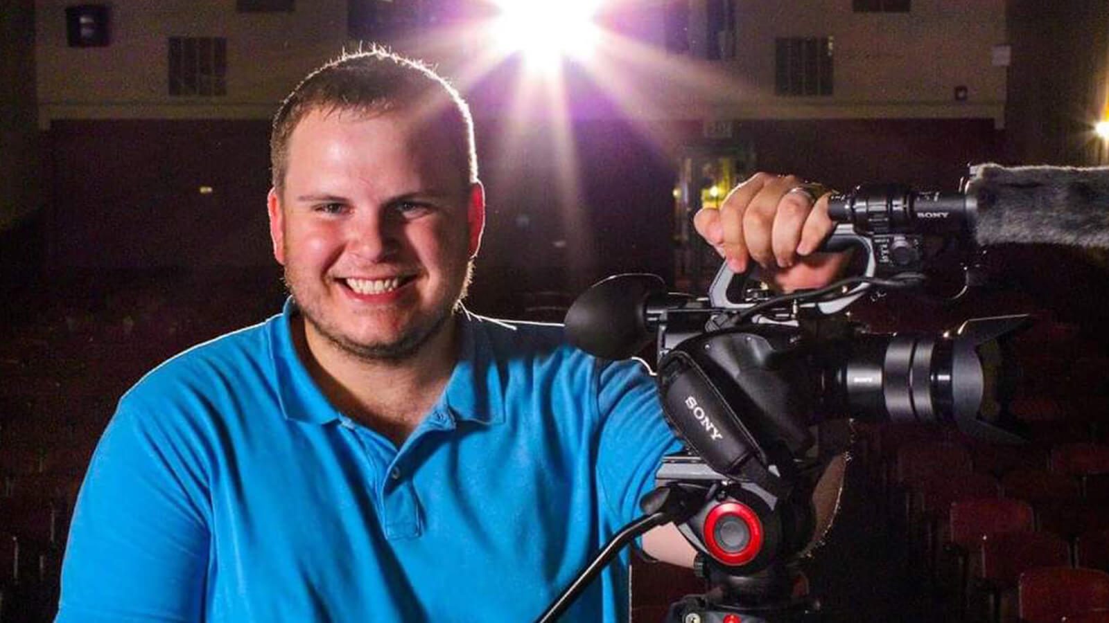 Grad Cody Grammer in a blue collared shirt, holding a video camera in a movie theater.