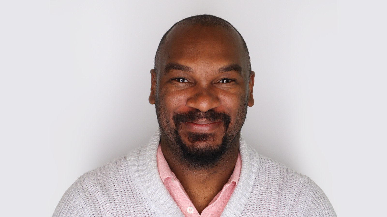 Quentin stands in front of a white wall wearing a white cardigan over a pink polo. His arms are crossed and he is smiling.