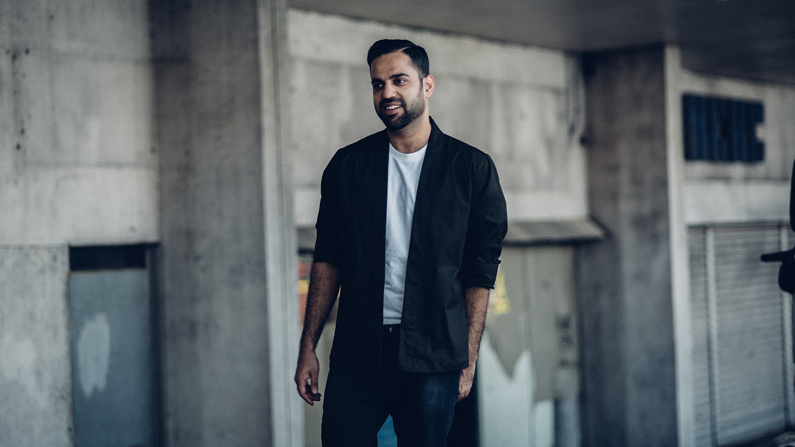 A well-dressed man in a white t-shirt and black blazer posing in front of a worn-down building.